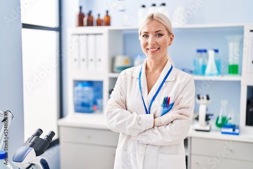 Young blonde woman wearing scientist uniform standing with arms crossed gesture at laboratory