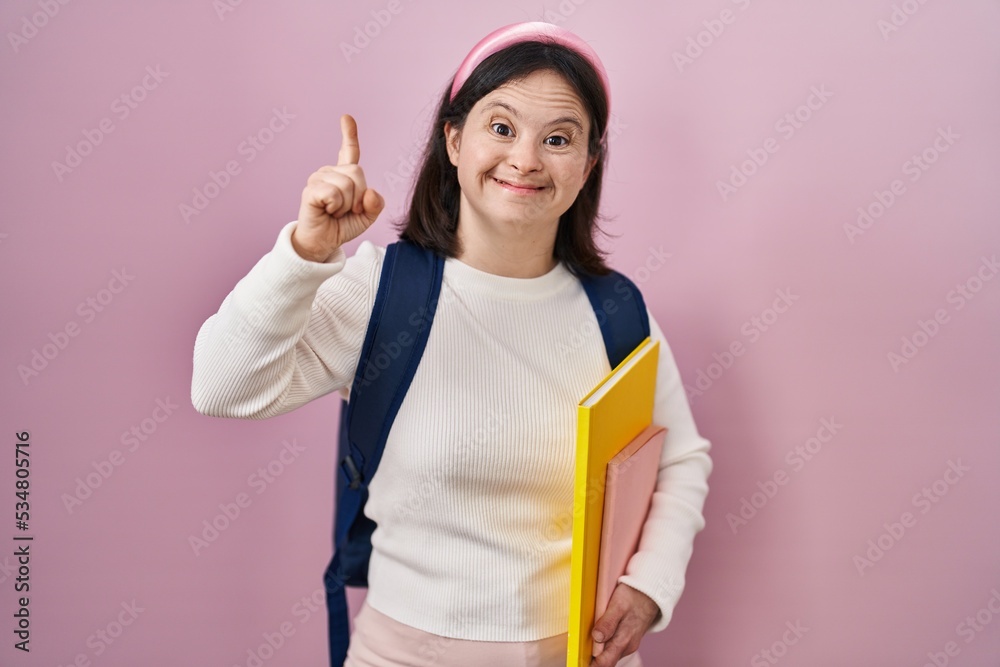 Woman with down syndrome wearing student backpack and holding books smiling amazed and surprised and pointing up with fingers and raised arms. Woman with down syndrome wearing student backpack and holding books smiling amazed and surprised and pointing up with fingers and raised arms.