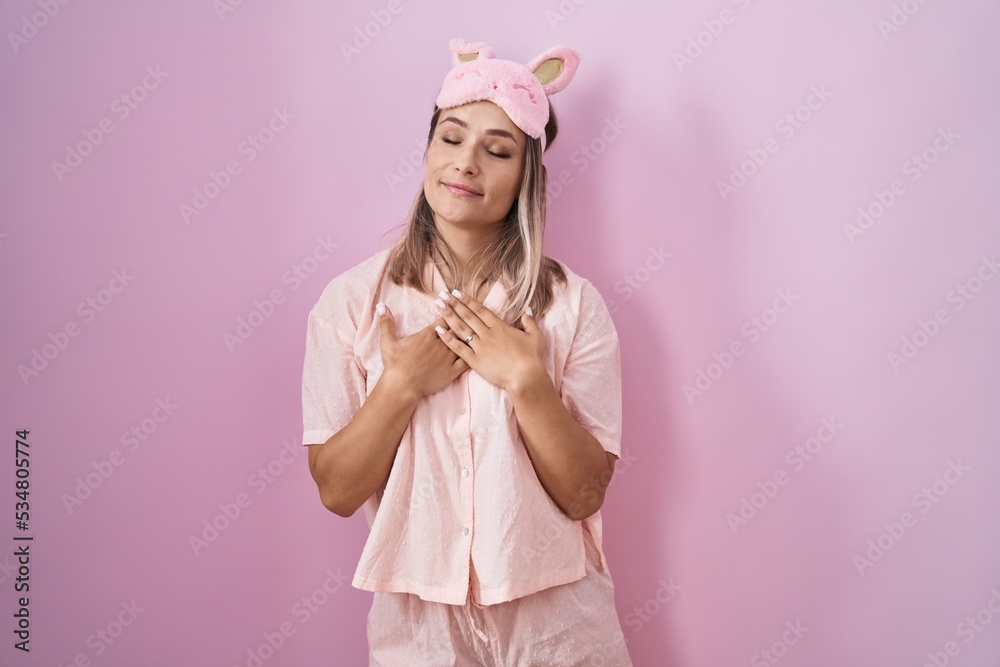 Blonde caucasian woman wearing sleep mask and pajama smiling with hands on chest with closed eyes and grateful gesture on face. health concept. Blonde caucasian woman wearing sleep mask and pajama smiling with hands on chest with closed eyes and grateful gesture on face. health concept.