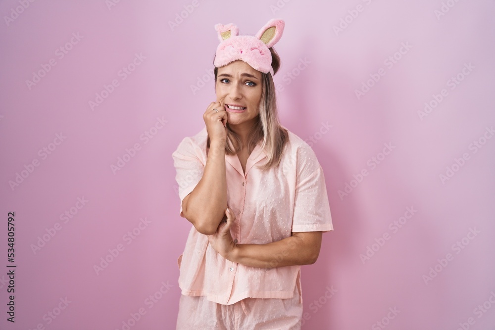Blonde caucasian woman wearing sleep mask and pajama looking stressed and nervous with hands on mouth biting nails. anxiety problem.