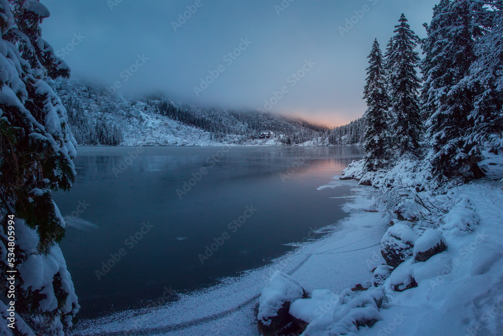 December winter sunrise in Tatra Mountains near lake. Thin ice covers the lake, fog go down the mountainside, fir trees covered snow. Houses are visible in the distance.