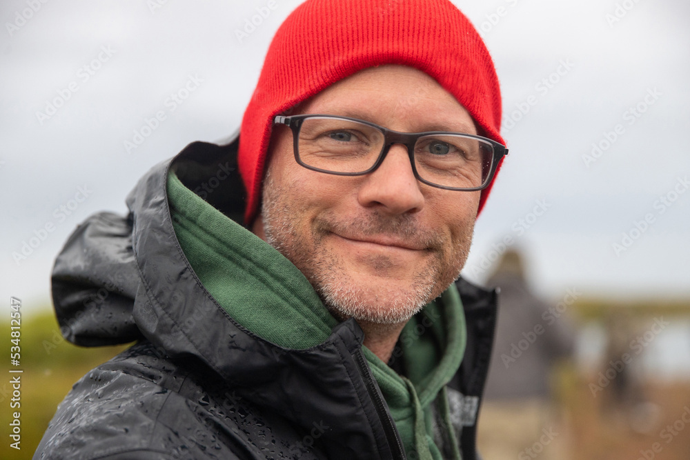 Close up face of handsome man wearing red beanie hat and glasses. He has beard stubble and a friendly smile. Close up face of handsome man wearing red beanie hat and glasses. He has beard stubble and a friendly smile.