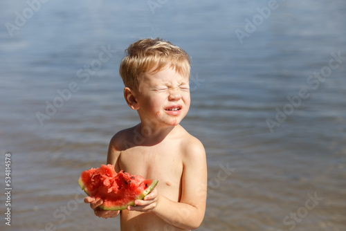 A blond boy 3-4 years old eats a piece of ripe red watermelon, looks at the bright sun, closes his eyes against the background of the river. On a sunny summer day