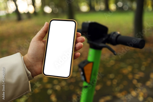 A girl stands with a scooter and shows a phone with empty white lighting in an autumn park