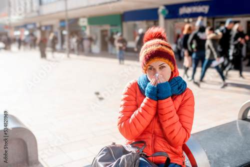 woman having a cold, wiping her nose and sitting against pharmasy in winter city 