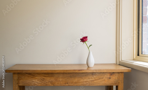 Single red rose in white vase on oak sidetable against beige wall next to window with sunlight