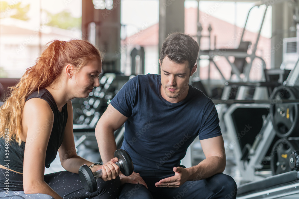 Young woman sitting on a stool with personal trainer, Trainer making fitness for female client at gym Young woman sitting on a stool with personal trainer, Trainer making fitness for female client at gym