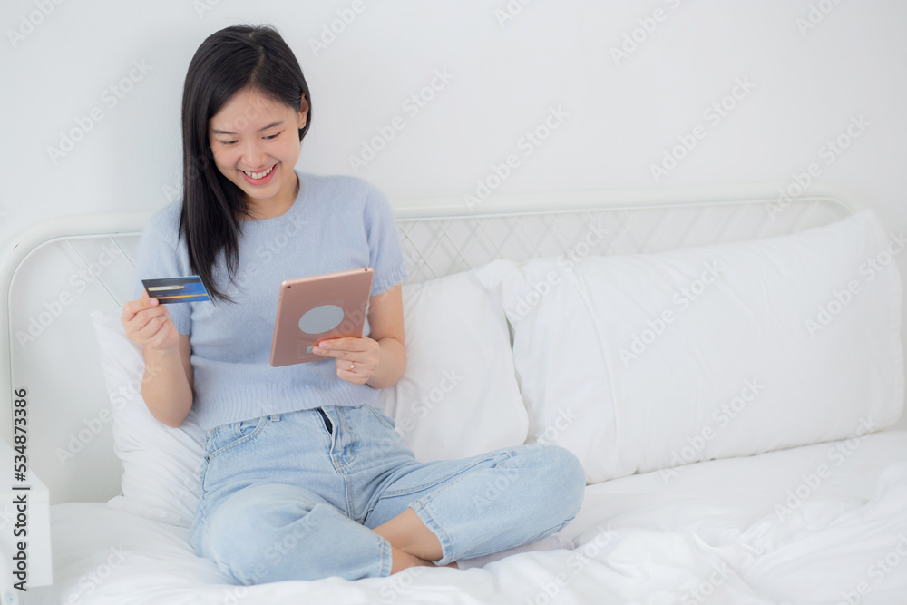 Young asian woman sitting on bed using digital tablet shopping online with credit card in the bedroom at home, female paying with transaction financial, purchase and payment, business concept. Young asian woman sitting on bed using digital tablet shopping online with credit card in the bedroom at home, female paying with transaction financial, purchase and payment, business concept.