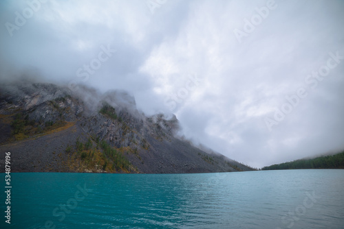 Dramatic meditate landscape with ripples on big turquoise mountain lake against high sharp rocks in low clouds. Shiny azure water of alpine lake and large rocky mountain range in low cloudy gray sky.