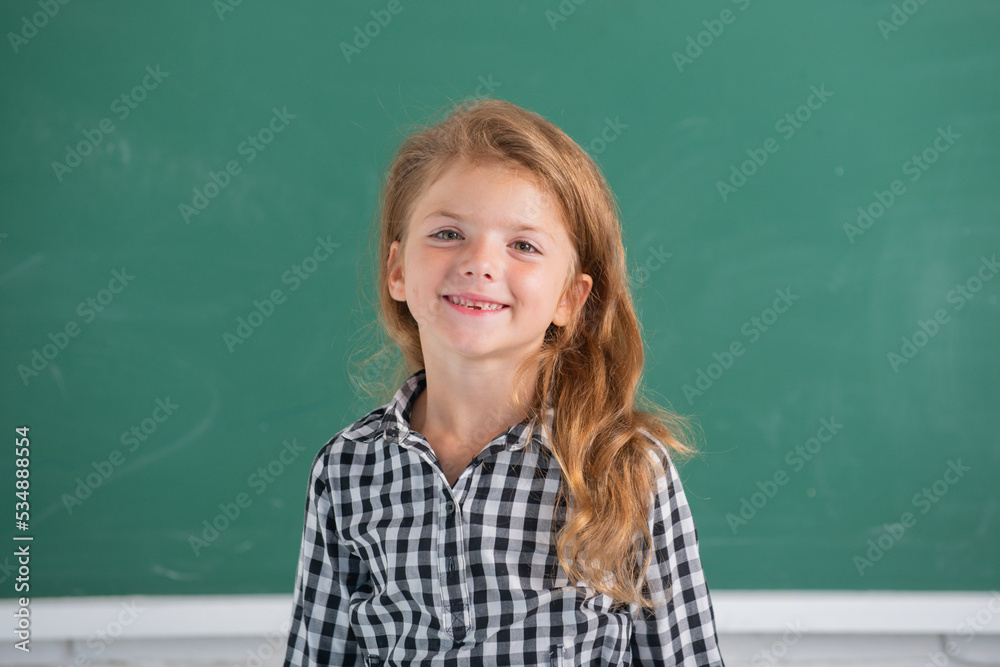 Close-up portrait of attractive small little cheerful girl sitting on table desktop in class room indoors. Little funny school girl face. Kids education and knowledge. Student kids.