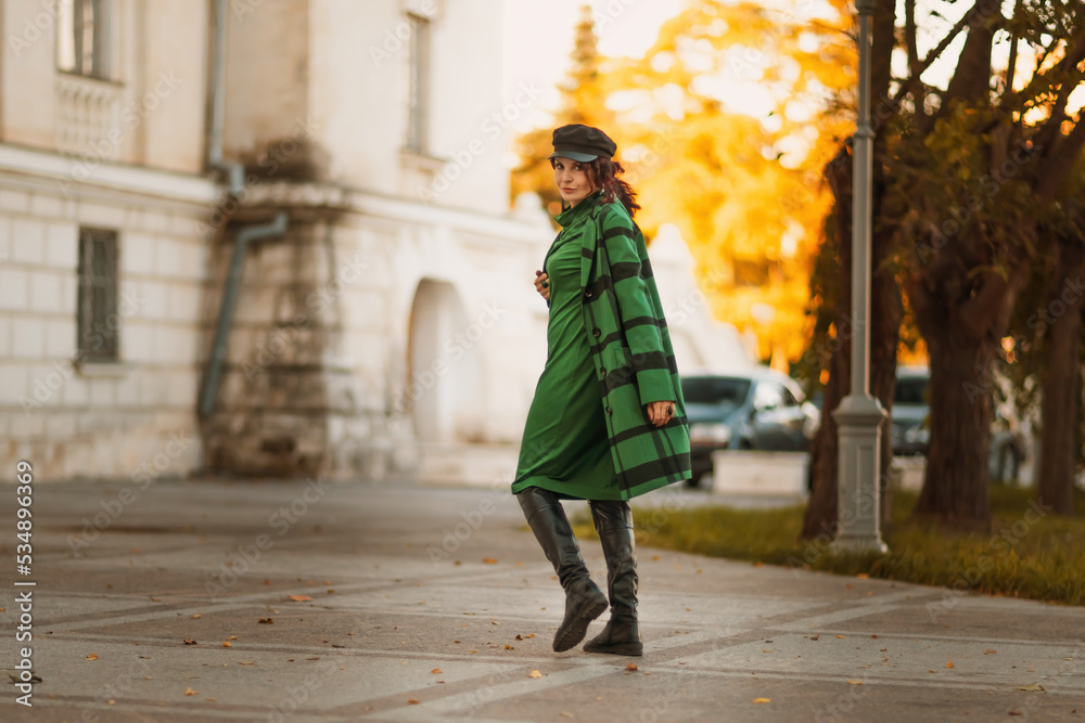 Outdoor fashion portrait of an elegant fashionable brunette woman, model in a stylish cap, green dress, posing at sunset in a European city in autumn.