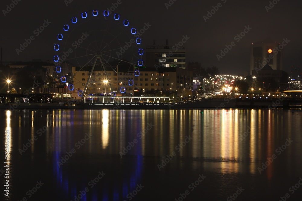 Night landscape with a view of the historical buildings of the city center, the Ferris wheel, evening lights and illuminations, as well as their perfect reflections in the mirror surface of the water.