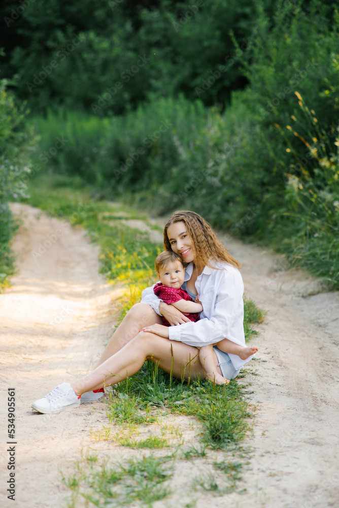 Naklejka premium Smiling mom holds her little son in her arms, sitting in the park on the path