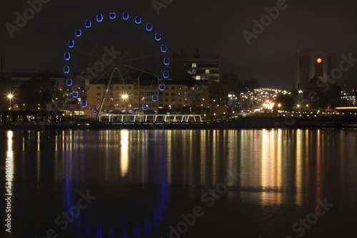Night landscape with a view of the historical buildings of the city center, the Ferris wheel, evening lights and illuminations, as well as their perfect reflections in the mirror surface of the water.