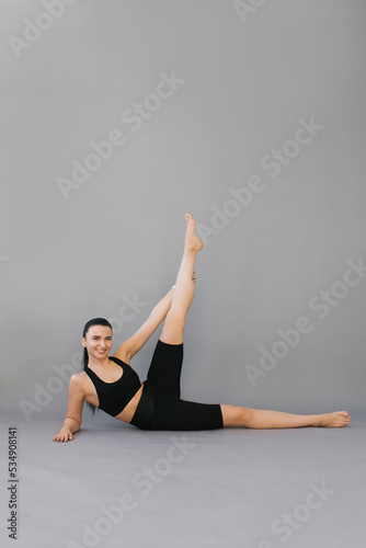 Woman does sports. A brunette in black sportswear is doing an exercise from yoga or fitness, she is lying on her side with her leg raised, isolated gray background