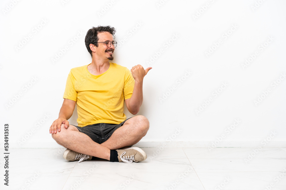 Young caucasian man sitting on the floor isolated on white background pointing to the side to present a product