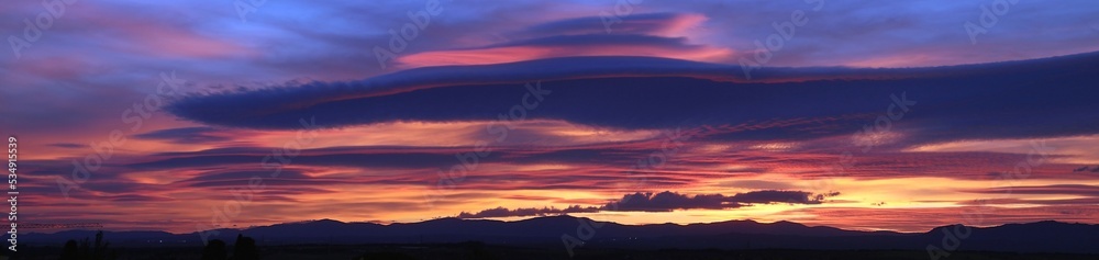 Atardecer en la Sierra de Guadarrama en Madrid, España. Cielo anaranjado y azul con una nube lenticular sobre la silueta de las montañas situadas al norte de Madrid.
