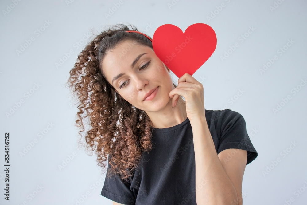 Picture of a lady holding a paper card with a heart with a thoughtful expression on her face, looking away for February 14, romantic date, black clothing, white background. Young curly haired woman. Picture of a lady holding a paper card with a heart with a thoughtful expression on her face, looking away for February 14, romantic date, black clothing, white background. Young curly haired woman.