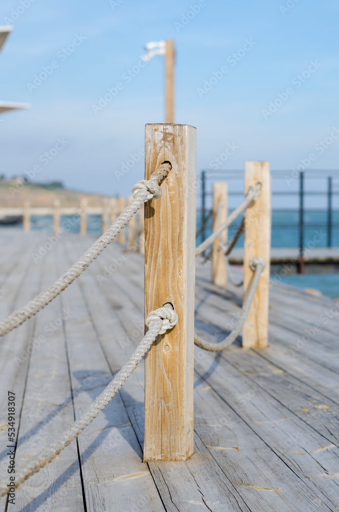 Fototapeta premium Wooden platform on the seashore. Fence made of old marine rope. Rope knot. Wooden poles. Yacht mooring.