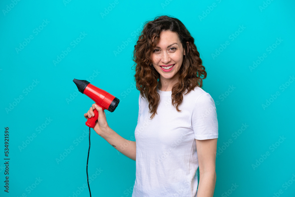 Young caucasian woman holding a hairdryer isolated on blue background smiling a lot Young caucasian woman holding a hairdryer isolated on blue background smiling a lot