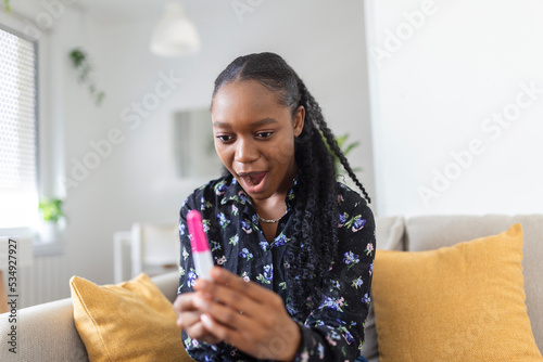 Young woman looking at pregnance test in happiness. Finally pregnant. Attractive black women looking at pregnancy test and smiling while sitting on the sofa at home photo