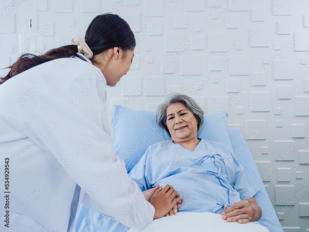 Happy Asian elderly senior woman patient in light blue dress lying on bed in saline solution was encouraging by young female doctor in white suit with stethoscope and holding hand in hospital room. Happy Asian elderly senior woman patient in light blue dress lying on bed in saline solution was encouraging by young female doctor in white suit with stethoscope and holding hand in hospital room.