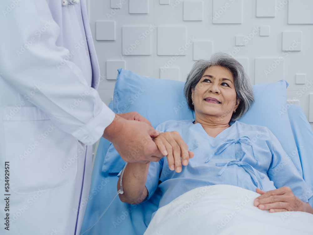 Happy smile beautiful Asian elderly old woman patient in light blue dress lying on bed while male doctor in white suit holding her hand and giving intravenous fluid on hand in the hospital room. Happy smile beautiful Asian elderly old woman patient in light blue dress lying on bed while male doctor in white suit holding her hand and giving intravenous fluid on hand in the hospital room.