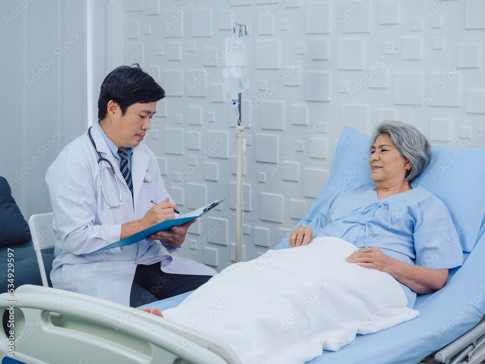Fototapeta premium Smiling Asian doctor in white suit take notes while discussing happy Asian elderly, senior woman patient who lying on bed with receiving saline solution in hospital. Health care and medical concept.