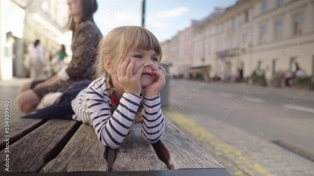 Little girl is gazes dreamily into the distance while lying on a bench on a street. The child rests in the city center. Family leisure in the city. Little girl is gazes dreamily into the distance while lying on a bench on a street. The child rests in the city center. Family leisure in the city.