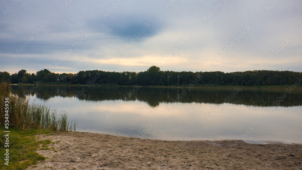 Fototapeta premium Ruhiger See mit Spiegelung des bewölkten Himmels am sandigen Ufer in der Dämmerung, Naturlandschaft am Gorinsee (Wandlitz), Landkreis Barnim, Brandenburg, Deutschland