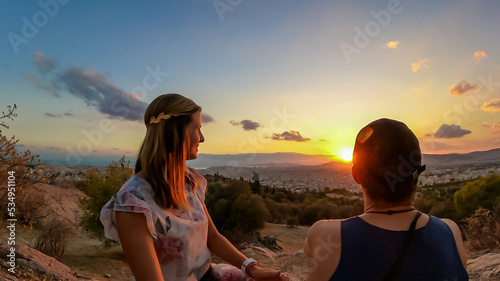 Young couple  watching sunset over city of Athens seen from Filopappou Hill (hill of muses), Athens, Attica, Greece, Europe. Athens cityscape and Aegean sea. Beautiful sunset point with aerial view photo