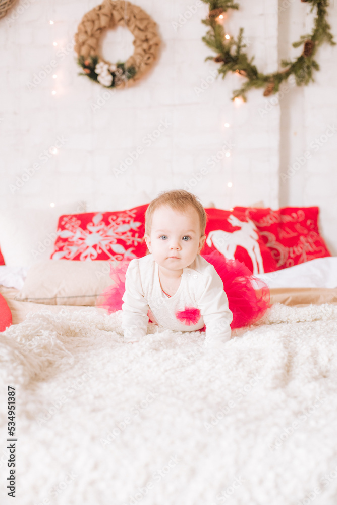 A little girl under one year old in an airy dress on a large bed in a room decorated for Christmas, among the pillows of garlands and pine needles. Christmas mood. Children and Christmas A little girl under one year old in an airy dress on a large bed in a room decorated for Christmas, among the pillows of garlands and pine needles. Christmas mood. Children and Christmas