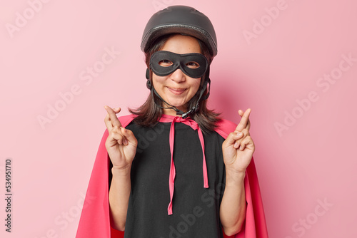 Horizontal shot of pleased brunette Asian woman keeps fingers crossed believes in good luck wears protective helmet eyemask and costume with cloak isolated over pink background comes on masquerade photo
