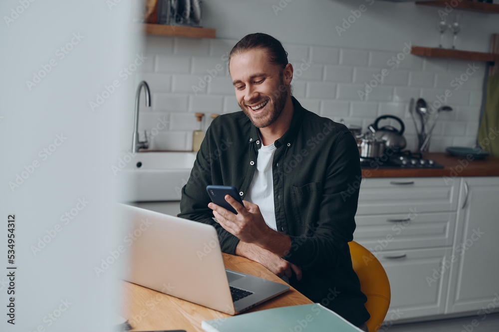 Cheerful young man using smart phone while working from domestic kitchen at home