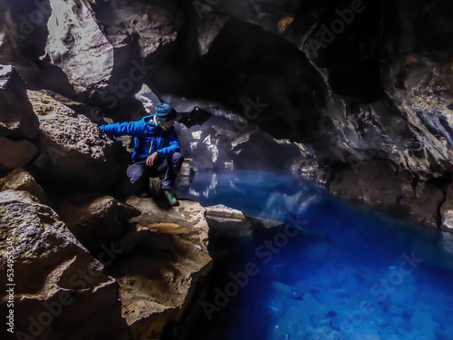 A young man wearing blue jacket squats at the rock in  Grj?tagj? Cave, looking down at the crystal blue water. Famous Game of thrones location. The water in this cave is full of minerals, it's hot pot photo