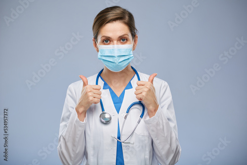 Smiling doctor woman with medical mask doing thumb up, isolated portrait.