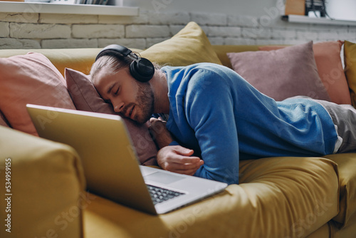 Young man in headphones sleeping near his laptop on the couch