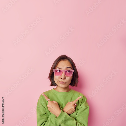 Confused young Asian woman with short dark hair points sideways with index fingers chooses between two options dressed in casual green jumper and trendy sunglasses isolated over rosy background © Wayhome Studio