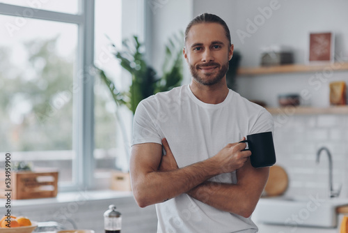 Handsome young man holding coffee cup and looking at camera while standing at the kitchen