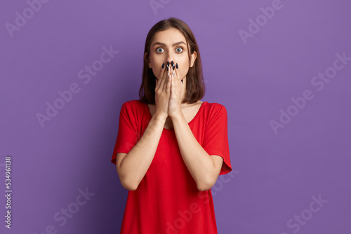 Its a crime. Call a police. Shocked young pretty brunette girl in red t-shirt covering her mouth with hands. Looking with horrified at the camera. Posing over purple background.