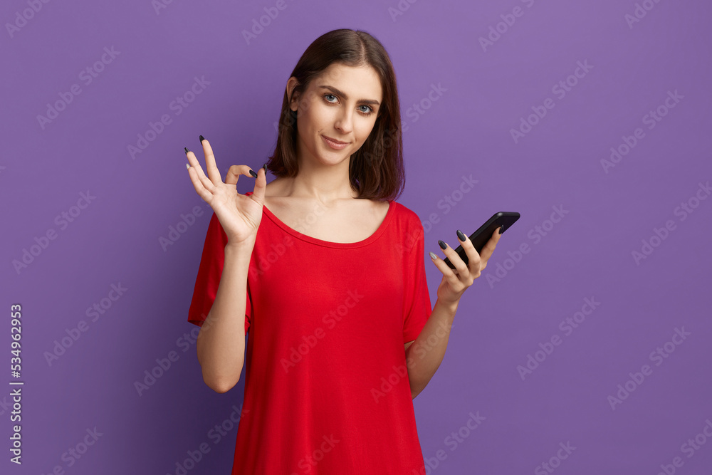 Digital life. Cheerful young pretty brunette girl in red t-shirt holds mobile phone and showing ok gesture. Using apps on her gadget. Using mobile banking for payment. Posing over purple wall. Digital life. Cheerful young pretty brunette girl in red t-shirt holds mobile phone and showing ok gesture. Using apps on her gadget. Using mobile banking for payment. Posing over purple wall.