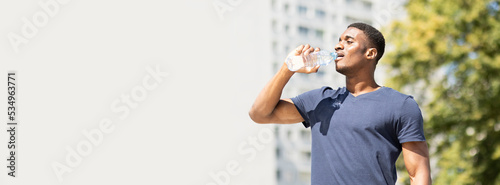 banner sportive young thirsty handsome african american man (guy) drinks water (isotonic) from plastic bottle after cardio workout outside, green tree and building are on background. Healthy lifestyle
