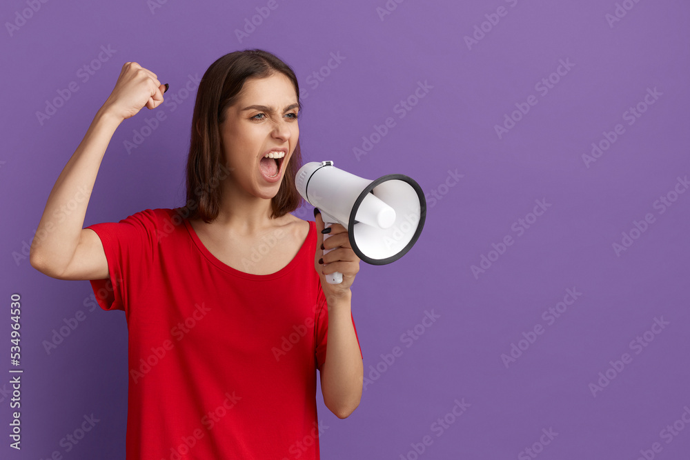 Naklejka premium Lets make the world great. We are against this policy. Confident pretty brunette girl in red t-shirt screams in megaphone. Protest for peace, calmness and equality. Feminism. Posing over purple wall.