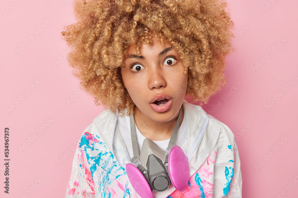 Shocked impressed young woman with curly bushy hair feels impressed keeps mouth opened cannot believe own eyes wears chemical protective clothing and antigas mask isolated over pink background