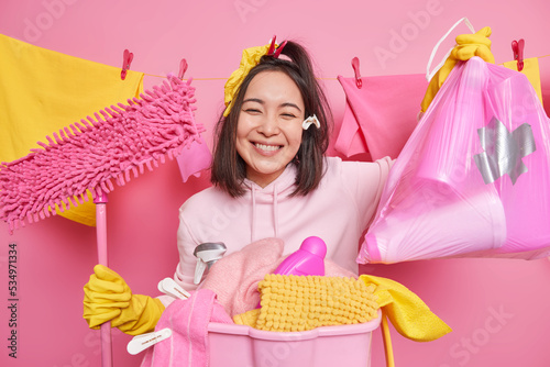 Regular cleaning concept. Happy dark haired housewife holds rubbish bag and mop for washing floor collects laundry in basket dressed in casual clothes poses against pink background washline behind photo