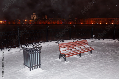 A red wooden bench with a black cast-iron urn on the embankment at night. Winter, snowfall