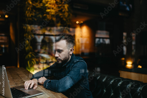 young male IT specialist with a beard and hairstyle works with a laptop in a cozy cafe. Online work on the Internet.