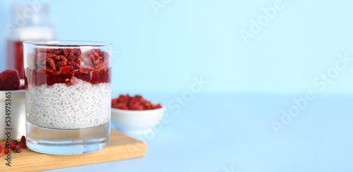 Glass of tasty pudding with goji berries and chia seeds on light blue background with space for text