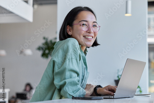 Pretty cheerful Asian woman in eyeglasses and casual clothes browses laptop computer connected to 4g internet updates software uses modern technologies poses in cafeteria looks gladfully away photo