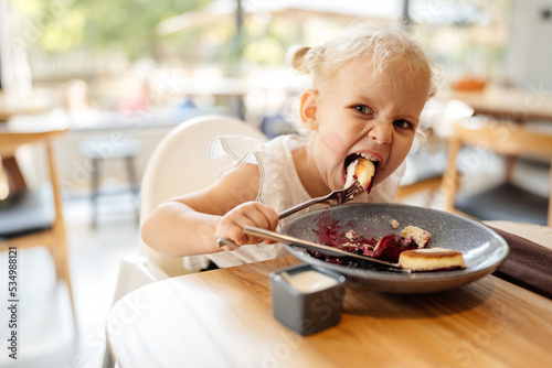 Fotografie, Tablou Hungry child girl blond curly with a fork in hand eats with a greedy look into the camera